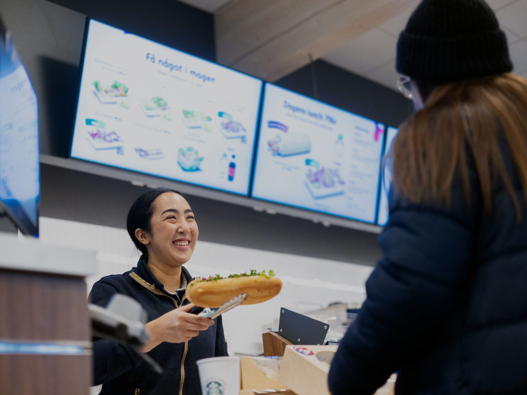 En kvinnlig butiksanställd håller fram en smörgås med serveringstång till en kund i förgrunden