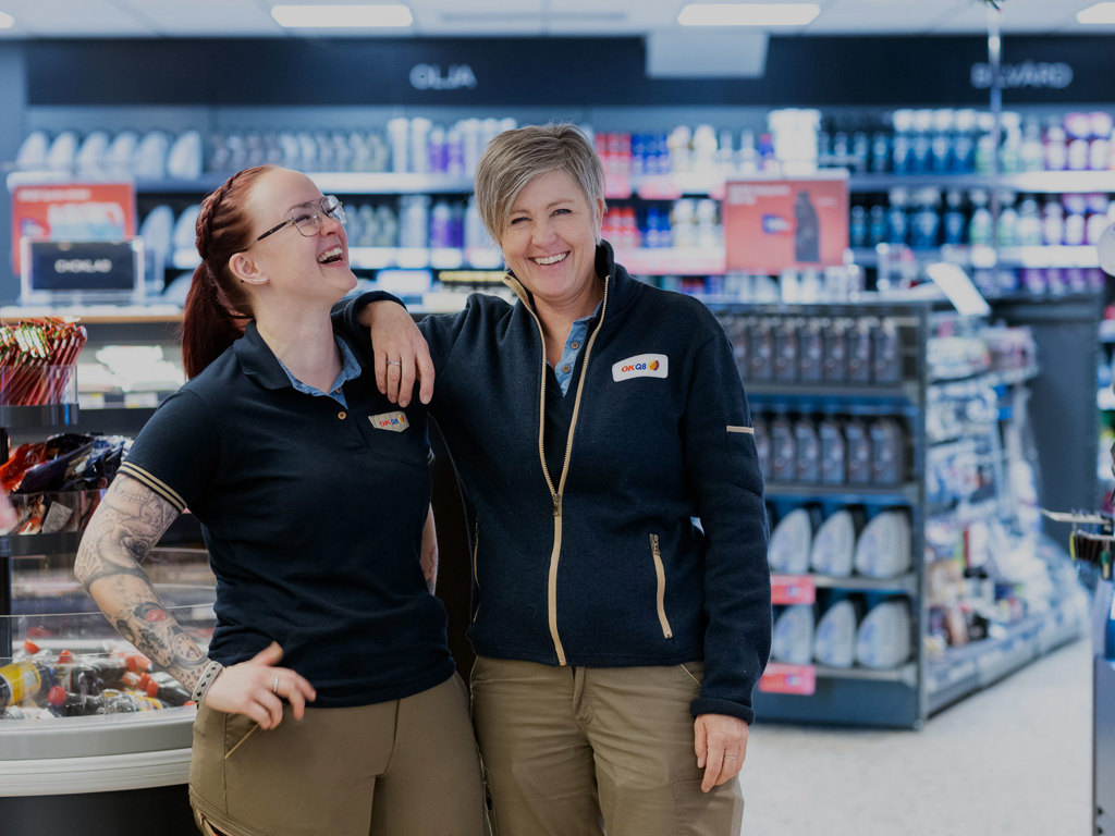 Två kvinnor i arbetskläder från OKQ8 skrattar i butiksmiljö